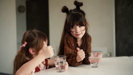 행복한 작은 다민족 자매들은 집 주방의 테이블에 앉아 순수한 미네랄 워터를 마시며 미소 짓고 좋은 징조를 보여줍니다.