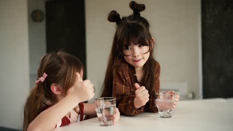 happy little multiracial sisters sit at table in home kitchen drink pure mineral water, smiling showing good sign