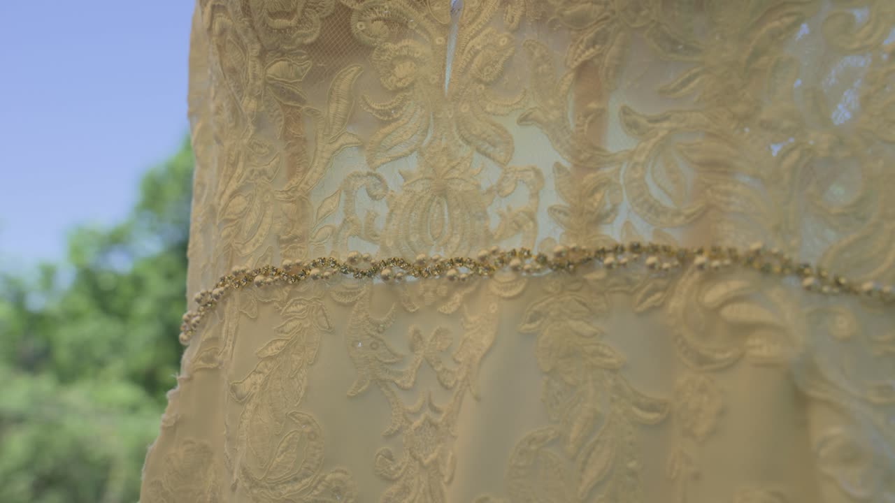 Une vidéo détaillée en gros plan présentant la ceinture scintillante en strass et la broderie délicate d'une robe de mariée, avec un panoramique révélant un arrière-plan flou d'arbres verts luxuriants et d'un ciel bleu.