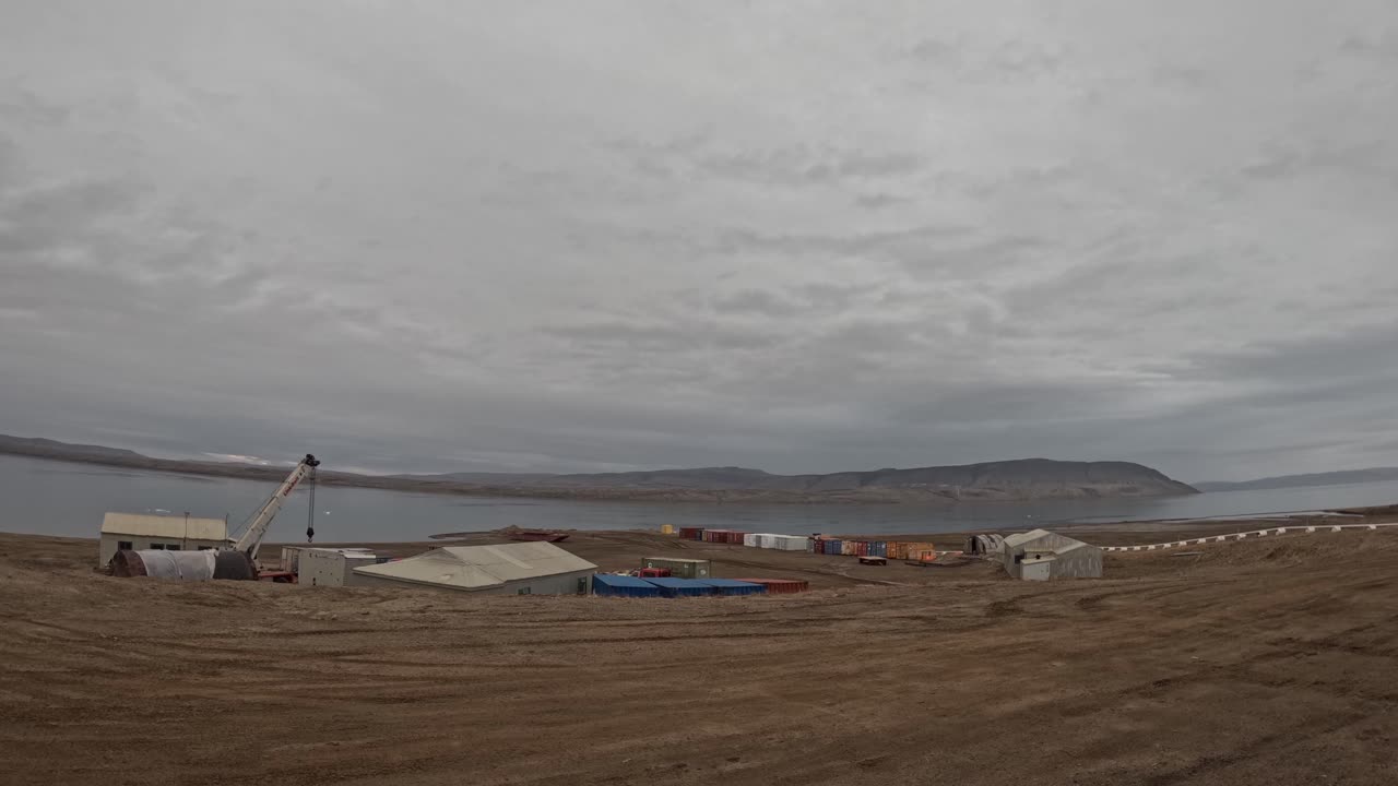 Timelapse overlooking research station buildings in Eureka, Nunavut, Canada. Arctic ocean bay and distant hills visible under calm summer conditions with moving clouds and soft natural light