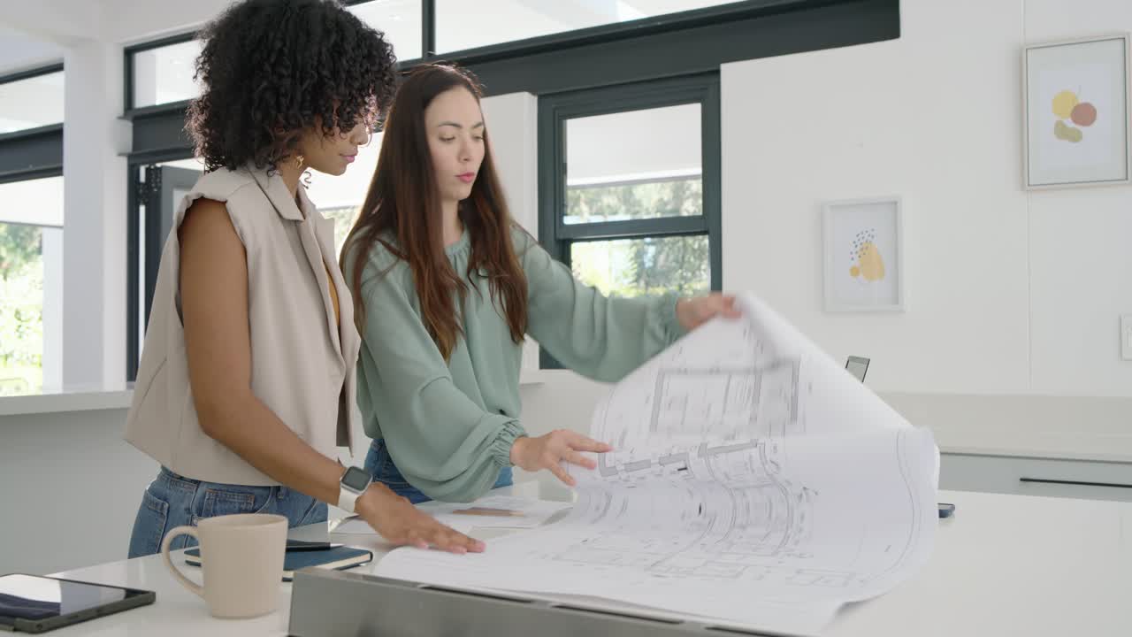 Mulheres diversas com colete bege e blusa menta espalhando e organizando planos na ilha da cozinha com um laptop para design