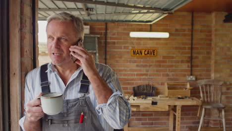 mature male wearing overalls using mobile phone in garage workshop with hot drink