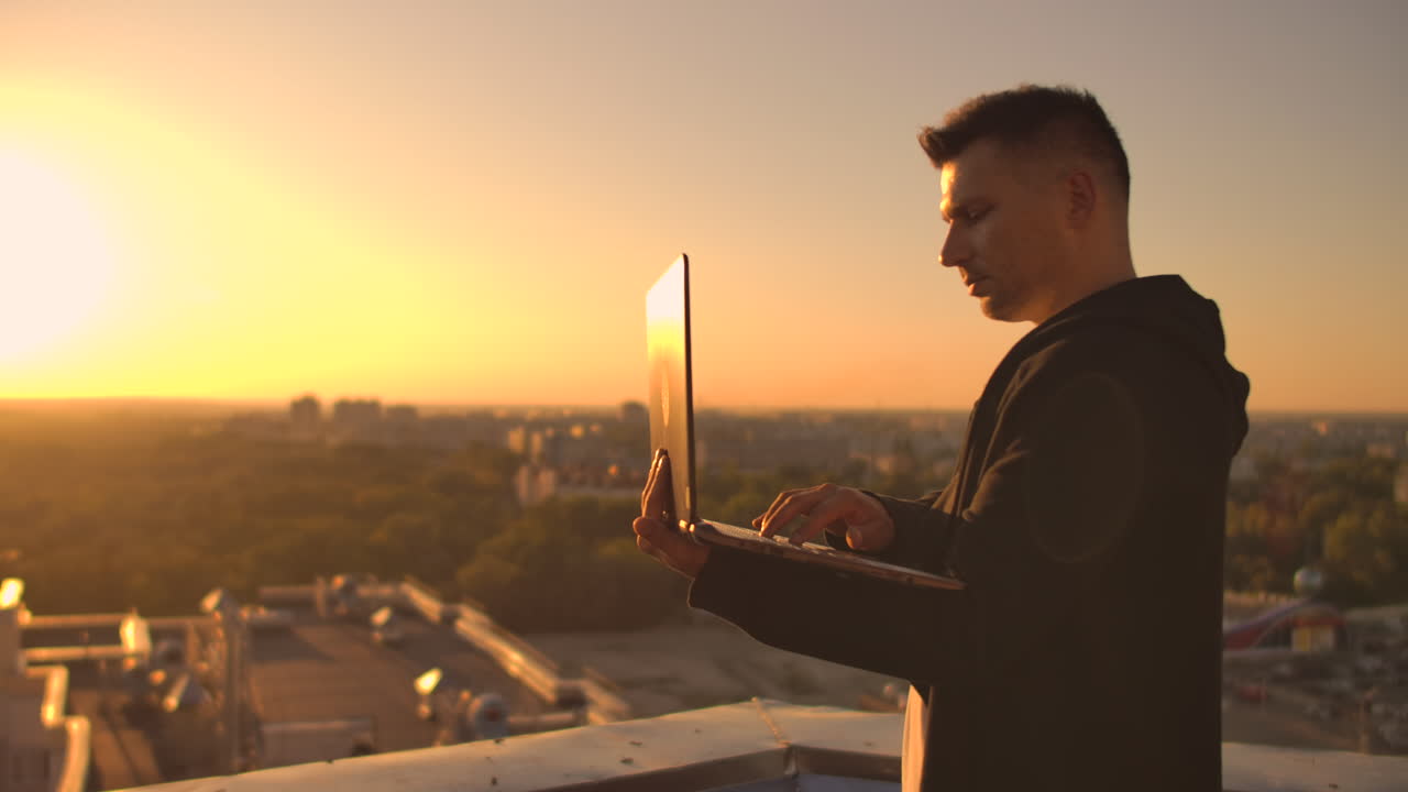 Hacker che usa il laptop sul tetto con vista sulla città e grafico forex. Concetto di hacking e statistiche. Un uomo al tramonto al rallentatore che scrive codice software su un laptop