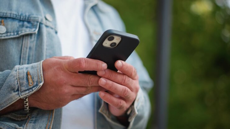 Primo piano di un uomo che tiene uno smartphone tra le mani. Sono visibili la texture della giacca di jeans, i dettagli delle cuciture, un bottone di metallo e un accessorio a catena al polso. Lo sfondo è leggermente sfocato.