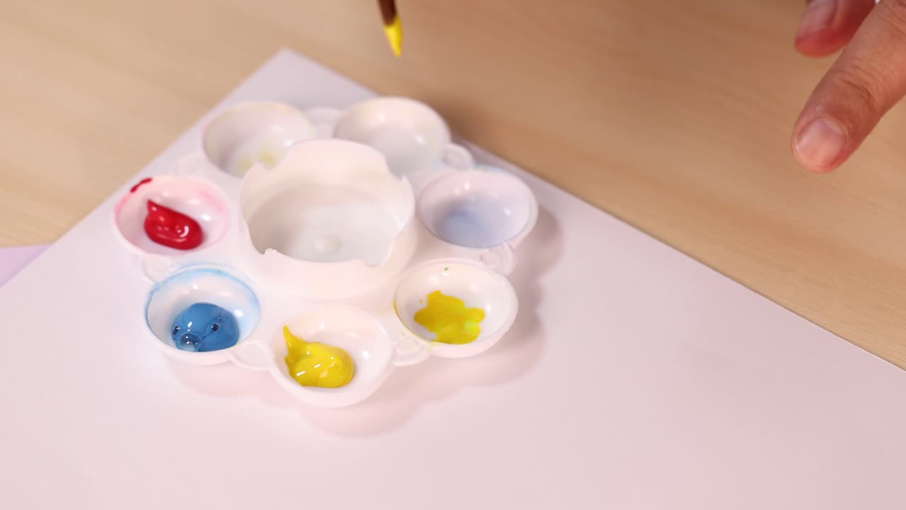 Artist hand uses brush to mix yellow watercolor paint in palette under soft indoor lighting