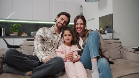 Portrait-of-a-happy-family,-a-brunette-man-with-stubble-along-with-his-brunette-wife-in-a-checkered-shirt-sitting-on-a-brown-sofa-along-with-their-little-daughter-in-a-pink-suit-in-a-modern-new-apartment