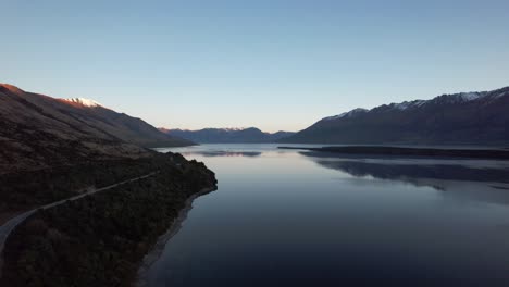 Aerial-flying-over-Lake-Wakatipu-with-the-road-next-to-it-and-surrounded-by-snow-capped-mountains-reflecting-in-the-lake-just-after-sunset-in-New-Zealand