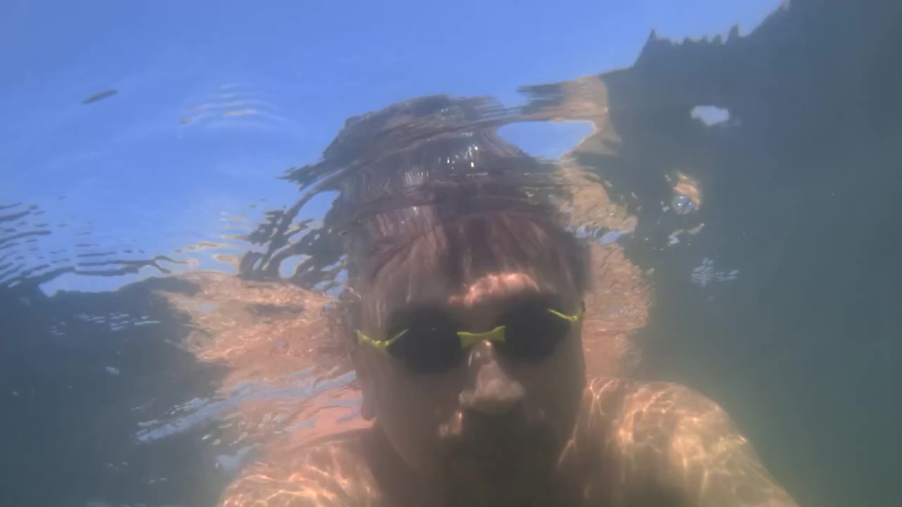 Man swimming underwater wearing yellow goggles with bubbles. Light rays from the surface are reflecting on his skin