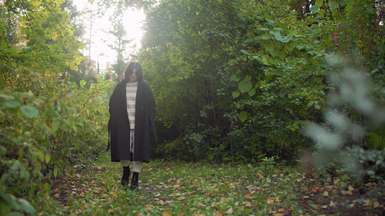 가을에 산책하는 사람이 재킷에 손을 넣고 고개를 숙인 채 숲길을 여유롭게 거닐고 있어요. 나뭇잎 사이로 부드러운 빛이 비치고, 차분한 걸음걸이, 긴 코트, 흰색 바지, 검은색 부츠, 푸른 잎, 발밑에는 낙엽이 흩어져 있어요.
