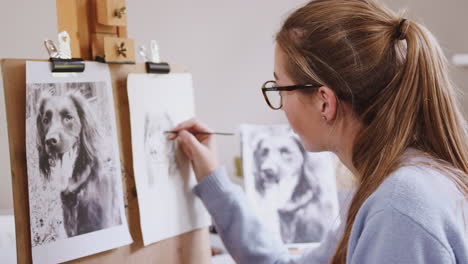 Female-Teenage-Artist-Sitting-At-Easel-Drawing-Picture-Of-Dog-From-Photograph-In-Charcoal