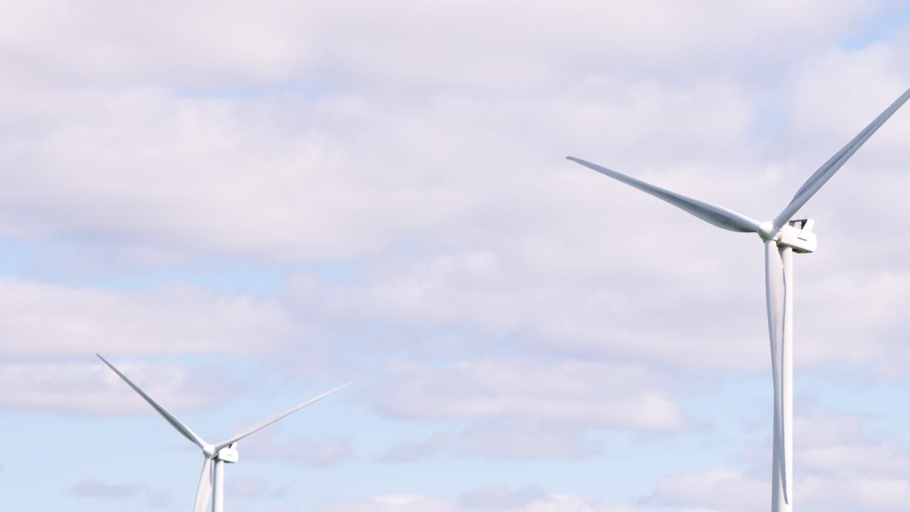 Large wind turbines slowly rotate under partly cloudy skies in a rural field near Edinburgh, Scotland, captured in a steady, wide shot with natural daylight