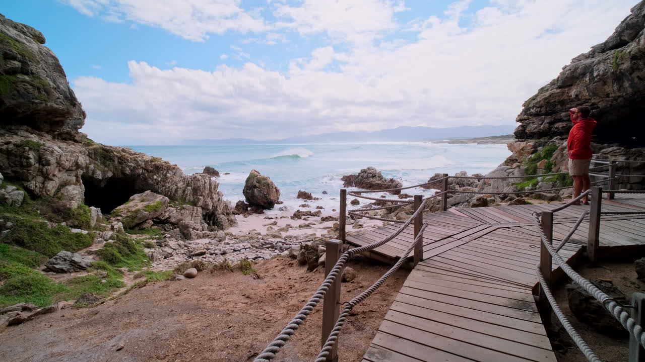 Male traveler on wooden boardwalk at historic Klipgat Cave looks out over ocean