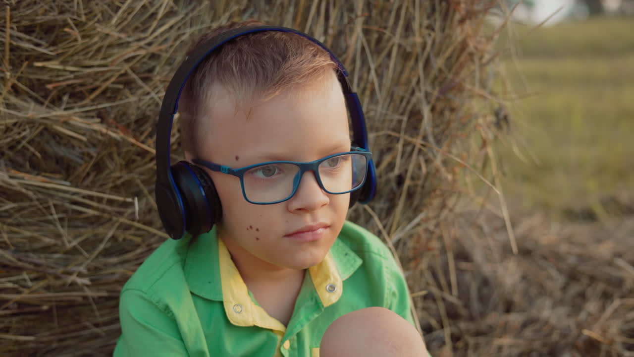 Criança pequena com fones de ouvido e óculos profundamente focada em ouvir música, sentada contra o feno em uma vasta área de cultivo ao pôr do sol, um momento sereno ao ar livre.