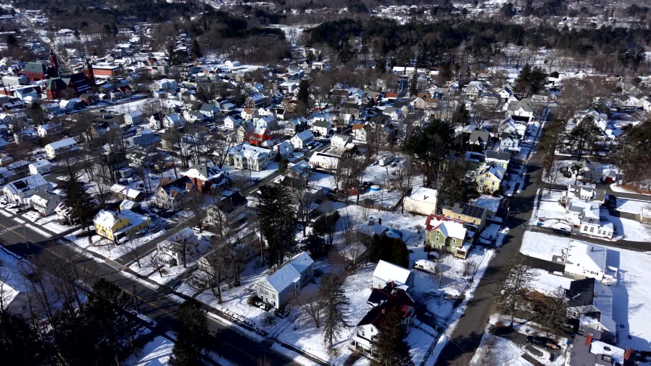 Vista aérea diurna de un pequeño pueblo americano cubierto de nieve fresca, mostrando casas residenciales, calles arboladas y una atmósfera invernal tranquila bajo la luz clara del sol. Vista cenital. Ware, MA.