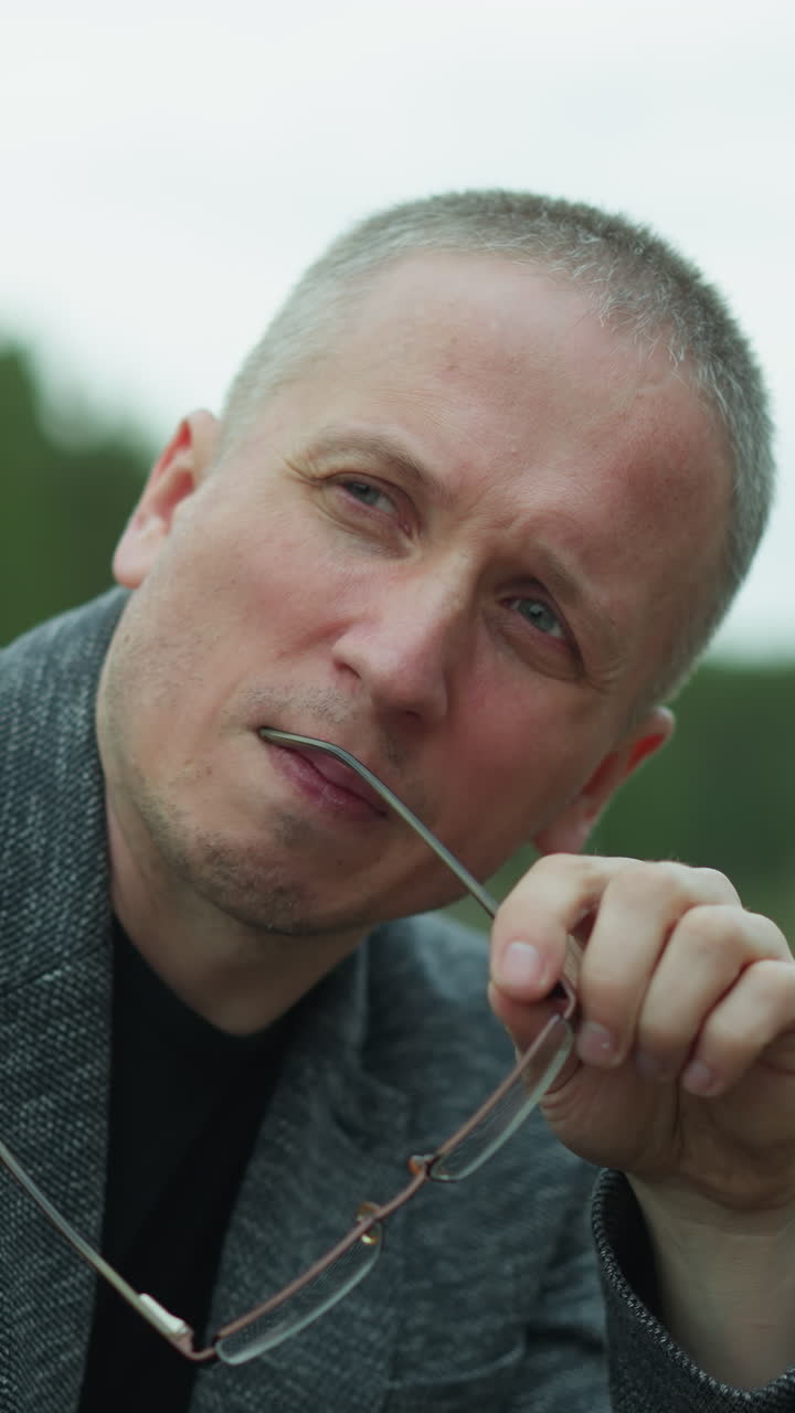 A close-up view of a middle-aged man adjusting his glasses while looking thoughtfully into the distance, he is outdoors, wearing a gray jacket, with a serene forest background