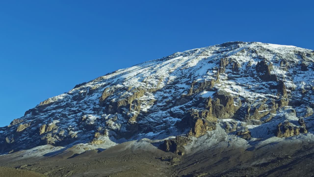 Ground-level view of Kilimanjaro shows its snow-covered rocky slope under a clear blue sky, capturing the dramatic volcanic terrain