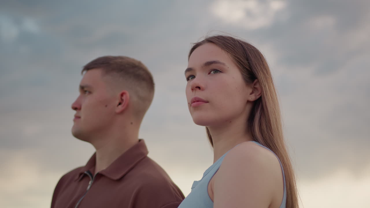 Young couple stands side by side beneath cloudy sky, man looking away with focus while woman gazes around with calm, introspective demeanor, both appearing reflective in soft evening light