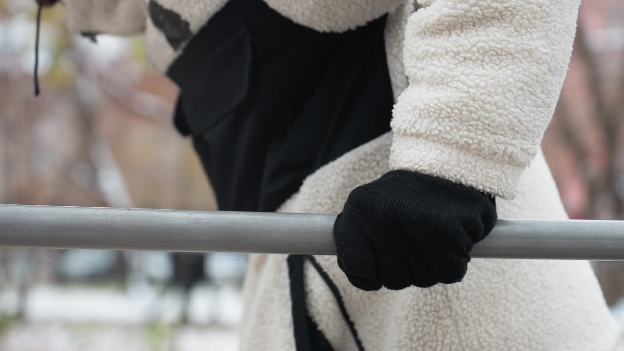 боковой вид человека в белой зимней куртке и черных перчатках, делающего подтяжки на открытом воздухе с размытым зимним фоном и далекими фигурами людей, гуляющих в городском парке во время холодной погоды