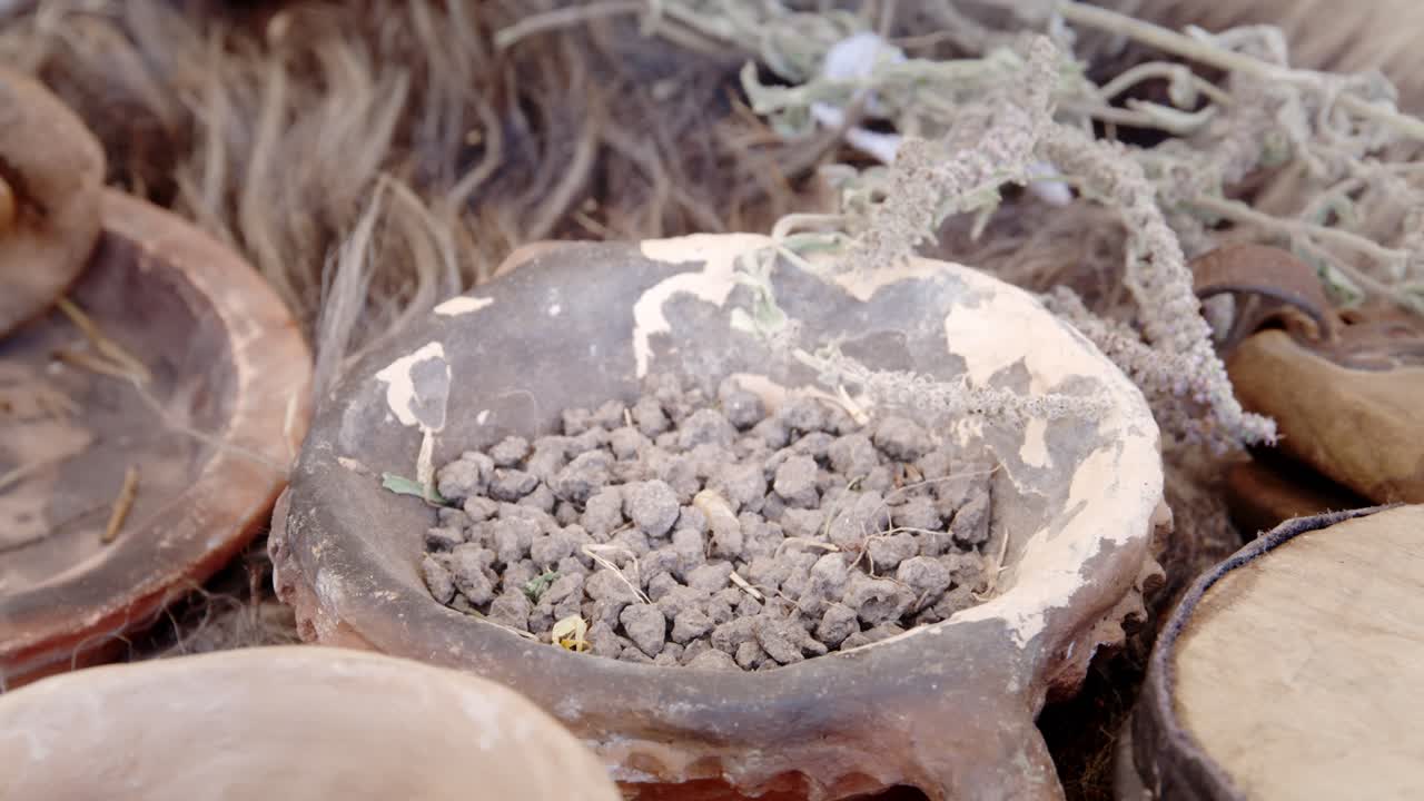 Extreme close-up shot shows a hand gently removing a piece of dry white sage or herb from a rustic clay bowl containing dark stones, emphasizing ancient and natural medicine