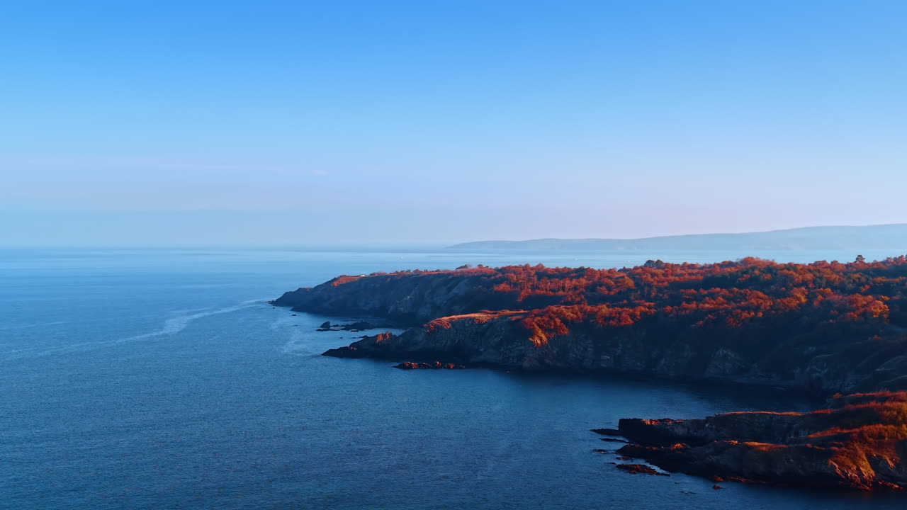 Klipper dækket med vegetation går over i det blå vandlandskab. Kystlinje med klipper ved havet eller oceanet ved solnedgang. Luftfoto