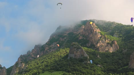 paragliding pilots fly paragliders among clouds and green mountains.