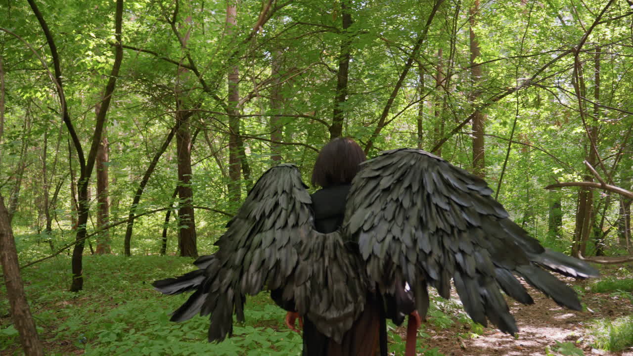 Back view of goddess with giant black wings walking through forbidden forest holding staff, surrounded by sunlight and shadows, symbolizing strength and divine elegance in magical green wilderness