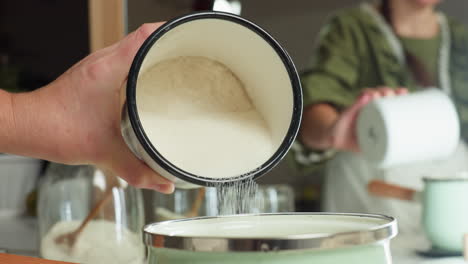 sugar being gently poured from enamel cup into green pot with stainless rim while kitchen mirror reflects person performing action, surrounded by glass jars