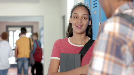 smiling teenage girl holding tablet and talking with friend in high school hallway