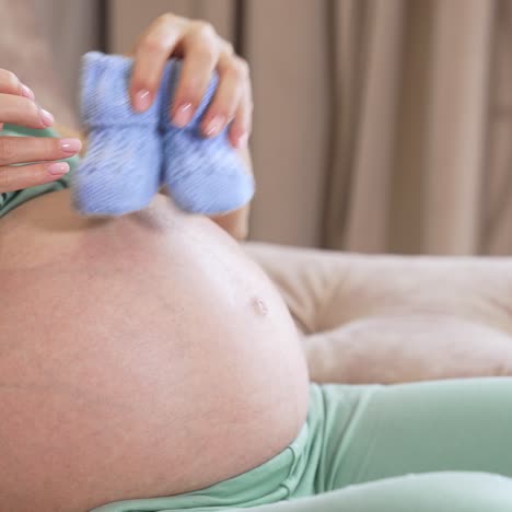 expectant mother is sitting on the sofa and playing with small baby socks on her big stomach. woman puts tiny baby booties on the armrest and sets her both hands on the belly to caress it