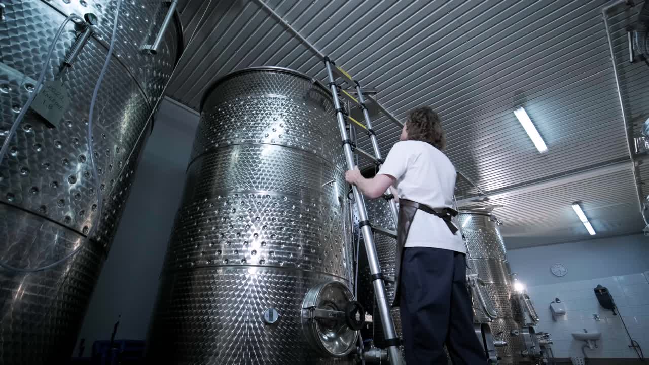 A happy successful farmer or winemaker is walking in the middle of wine tanks and checking a fermentation grade of high quality wine production in a modern winemaking facility or winery.