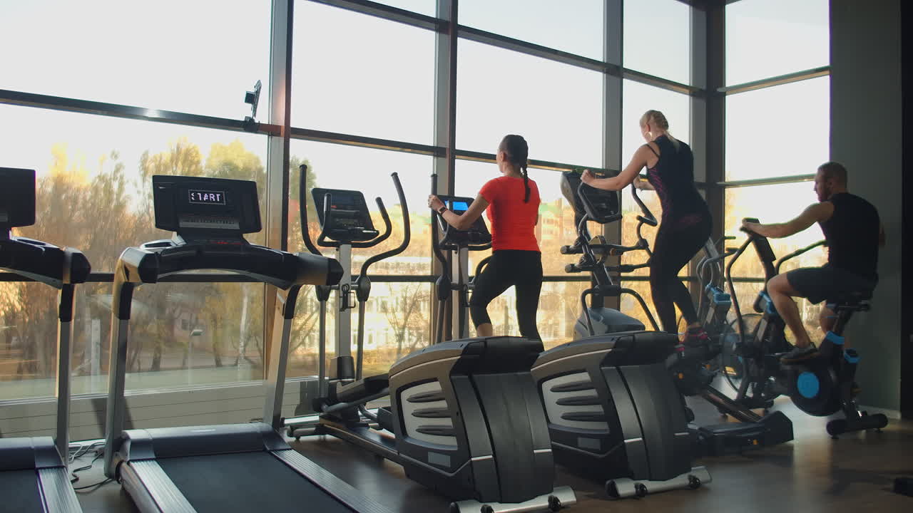 Young fit woman using an elliptic trainer in a fitness center. A group of young women train on sports training equipment in a fitness gym. Steady cam shot.