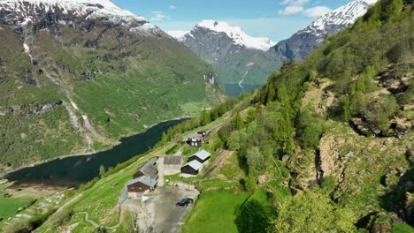 impresionante fiordo de geiranger revela desde la empinada ladera y la granja local - adelante moviéndose desde el aire en un paisaje verde exuberante con montañas cubiertas de nieve en el fondo - noruega