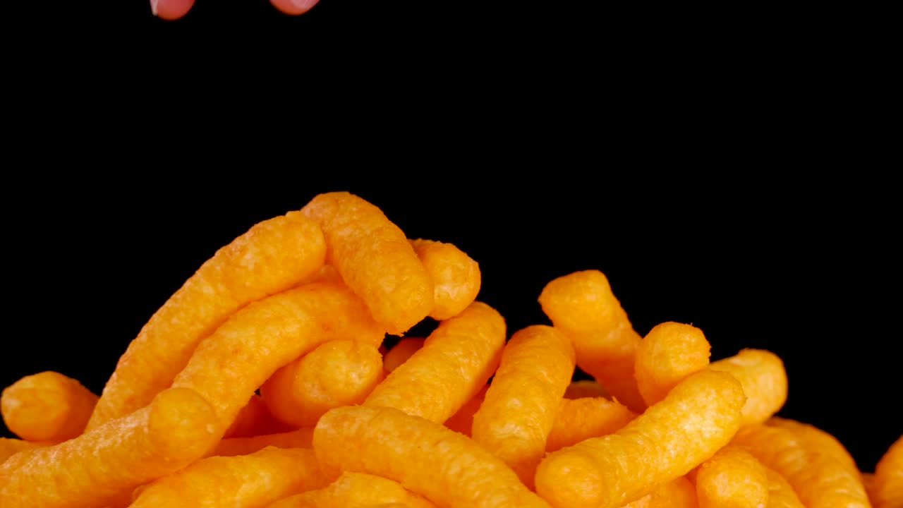 Human hand selects and lifts cheese puff snack from pile, brightly lit, black background, close-up