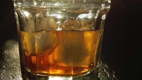 GLASS-WITH-ICE-CUBES-BEING-FILLED-WITH-LIQUOR-OR-WHISKEY-COLOR-AMBER-LIGHTED-FROM-BACK