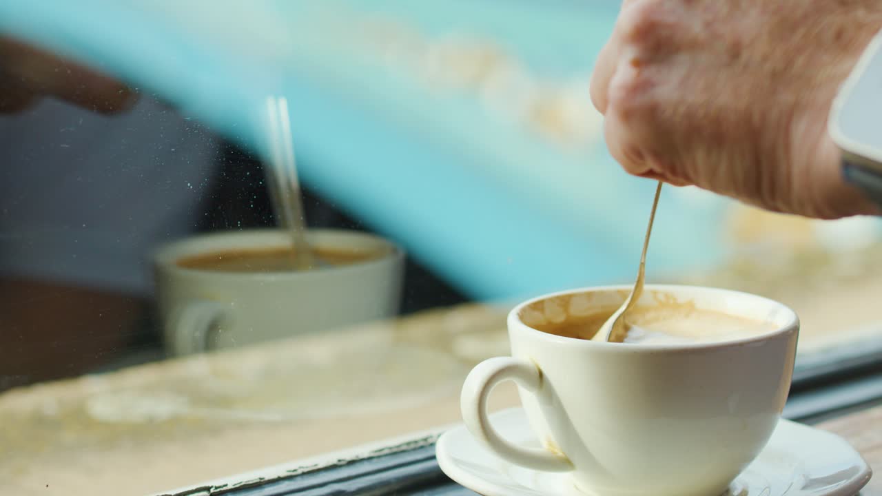 Hand stirs coffee in white cup at cafe table, natural daylight, close-up, steady camera