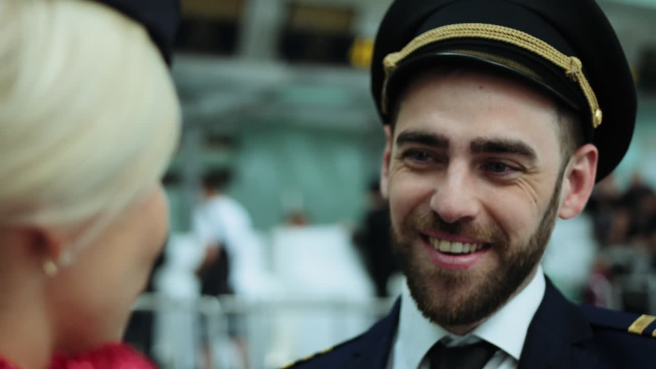 Hostess aeroportuale con capelli biondi che sorride affabilmente, parlando con il pilota. Donna europea vestita con un'uniforme blu scuro, una sciarpa rosso scarlatto annodata sulla sua camicia bianca. Ripresa cinematografica con telecamera RED. 2 modelli.