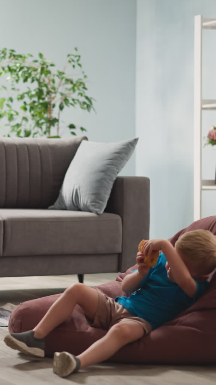 Little girl turns gymnastic ring hula-hoop toddler boy eats bun in bean chair on floor slow motion. Children rest in living room at home. Activity and laziness