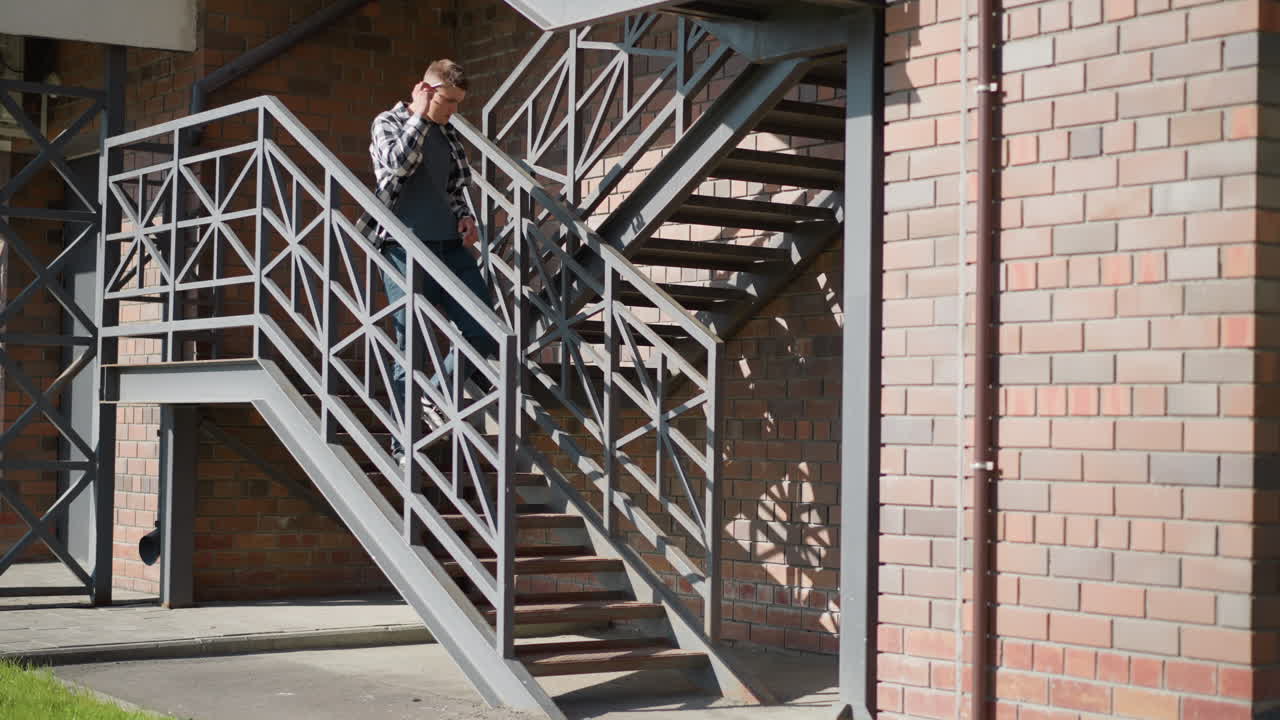 white boy descending exterior metal staircase pauses placing cigarette between lips lifts lighter flicks flame to ignite tip ember glows as grass and foliage sway gently in breeze beside brick wall