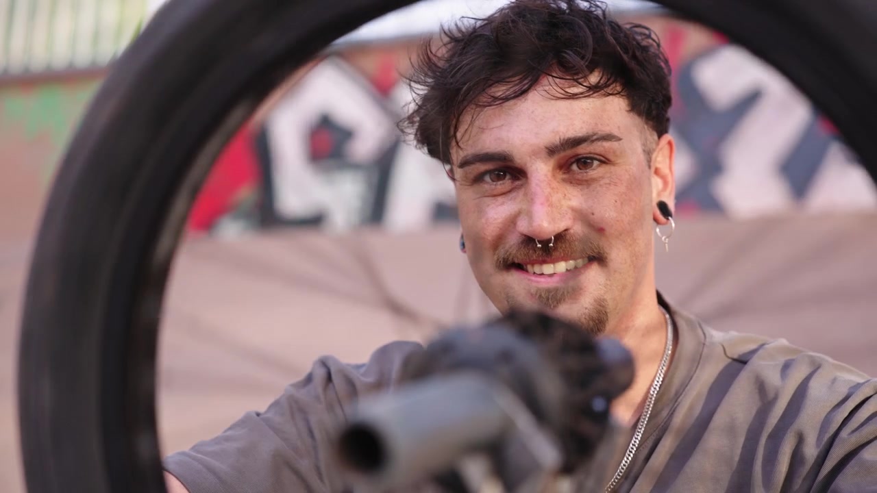 Porträt eines lächelnden BMX-Fahrers, der in einem Skatepark sein Vorderrad nah an der Kamera in Zeitlupe dreht. Urbaner Lifestyle und Action-Detail.