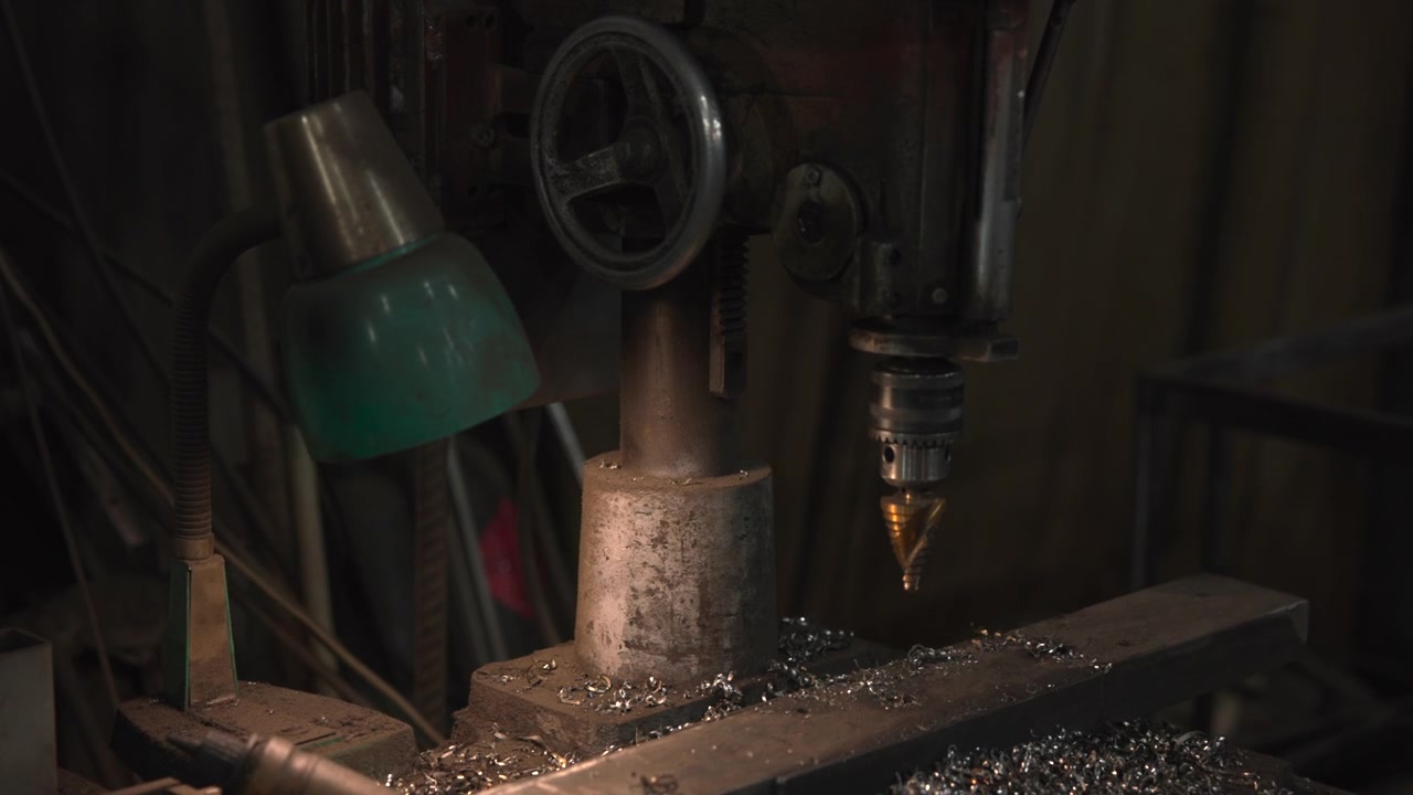 Postazione di lavoro industriale per la metallurgia in un'officina poco illuminata, con mandrino del trapano, punta da taglio a gradini, volantino usurato, morsa impolverata, trucioli arricciati, cavi aggrovigliati e superfici ruvide e arrugginite su un banco da lavoro disordinato.