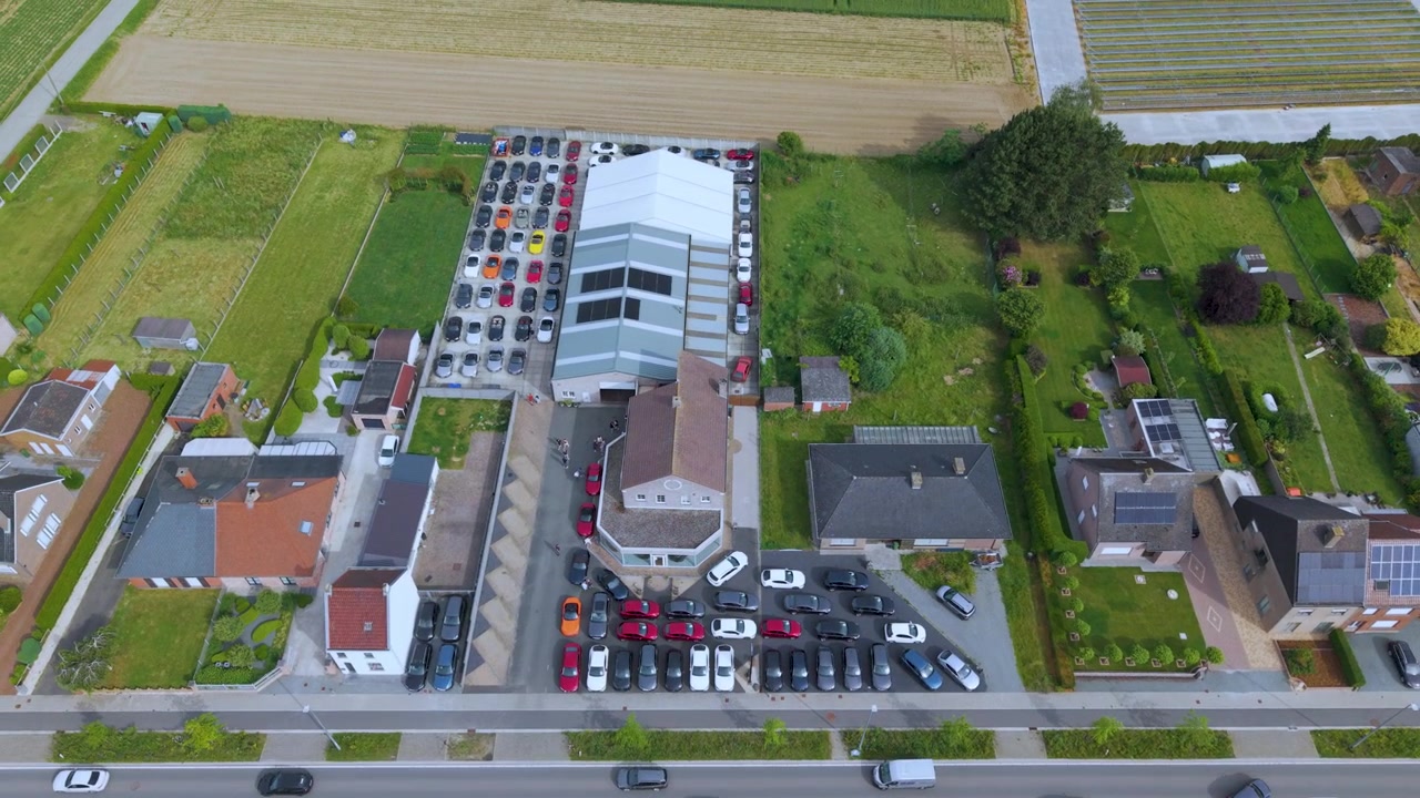 Vertikal luftoptagelse, der stiger op over en erhvervsbygning og et organiseret parkeringsområde. Optagelsen afslører nærliggende gader, boligblokke, åbne marker og et klart dagslandskab.