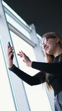 beautiful woman doing photo of herself indoors. attractive girl in glasses stands near the large windows and using mobile phone. view from below. vertical video