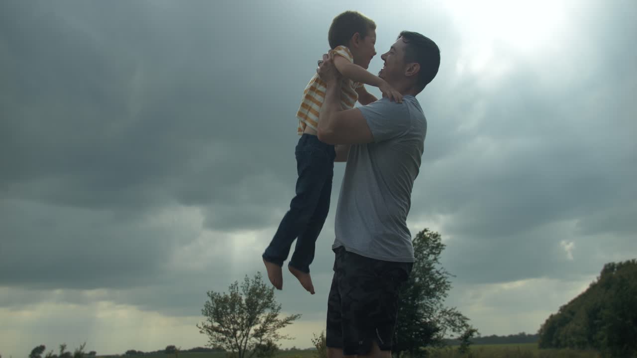 A happy, loving father holding and hugging his son for a warm, positive family moment outside under the sunlight shines through clouds.