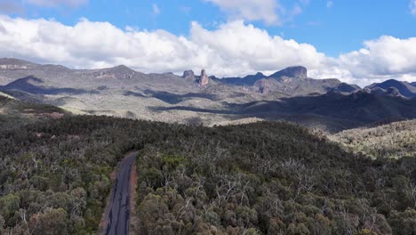 drone camera glides above dense eucalyptus forest toward rugged mountain range under bright daylight, revealing expansive australian wilderness and winding road below