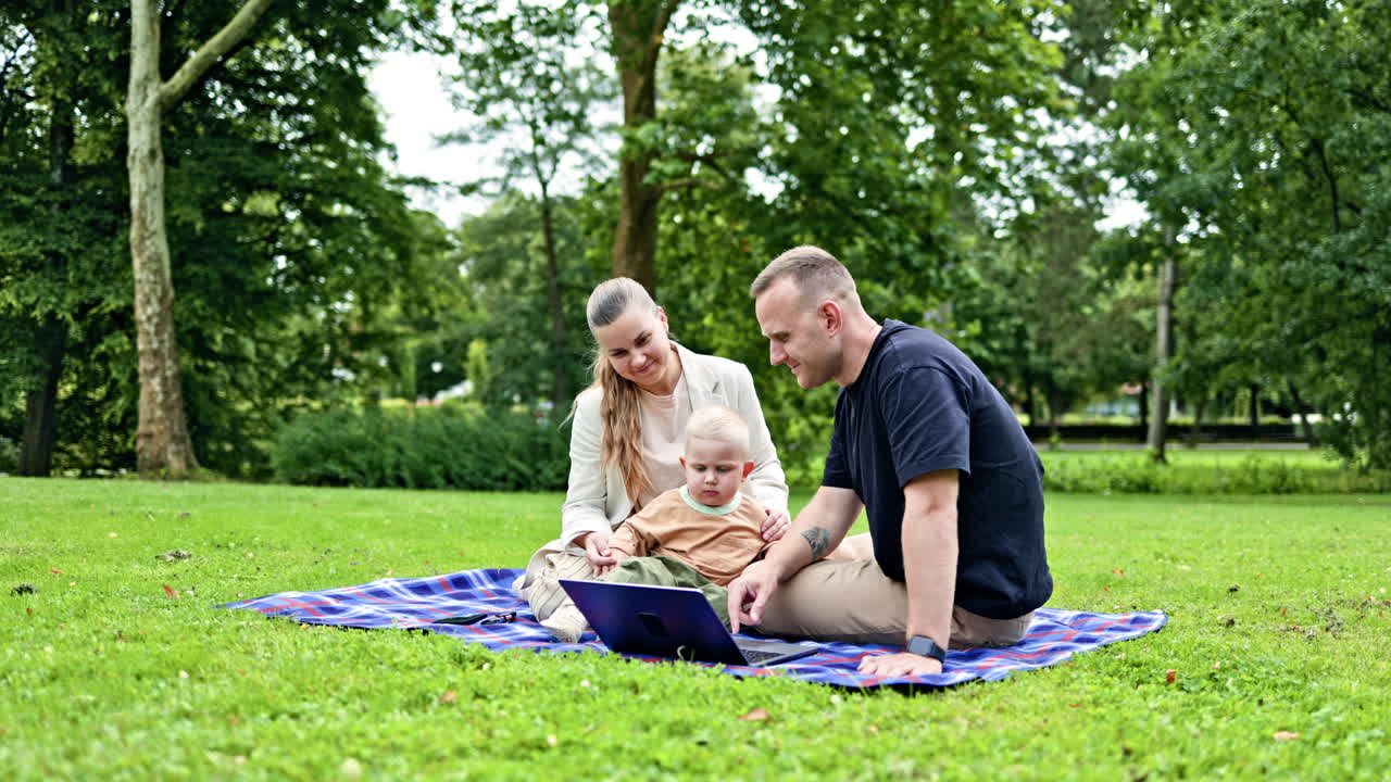 Glad ung familie med barn udendørs bruger bærbar computer og smartphone. Forretningsfamilie med lille barn arbejder fjernstyret, mens de går med barnet. Freelancefamilie med barn i parken.