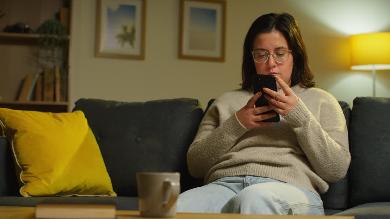 Young Woman Spending Evening At Home Sitting On Sofa With Mobile Phone Scrolling Through Internet Or Social Media 4