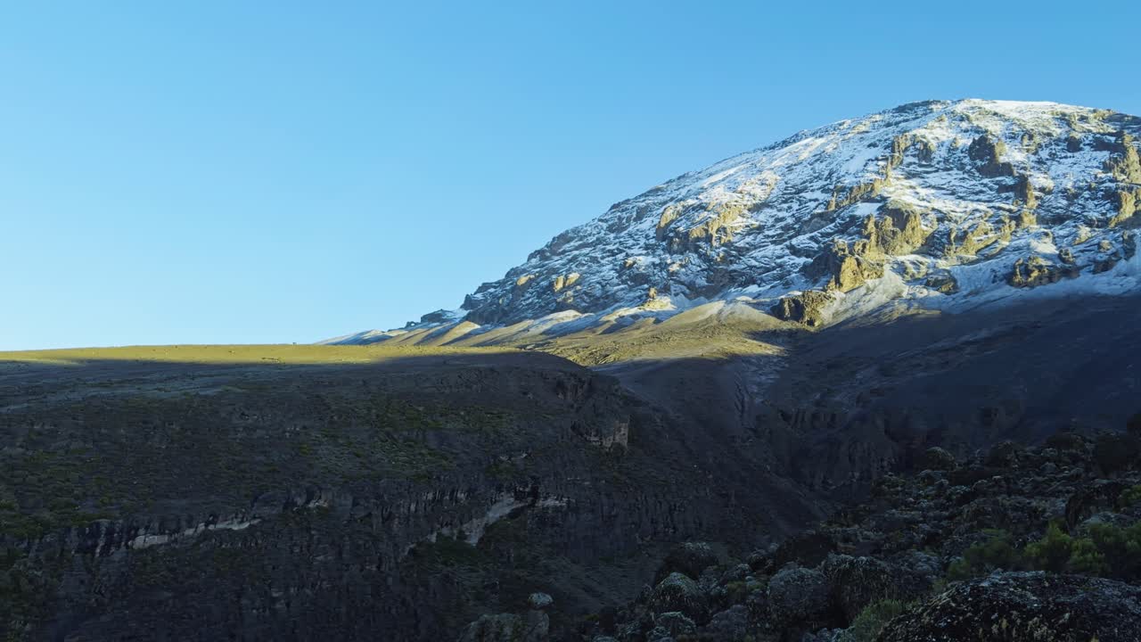 Ground-level shot captures a hiker raising arms in triumph at the rocky base of snow-capped Mount Kilimanjaro, highlighting the rugged terrain and clear blue sky