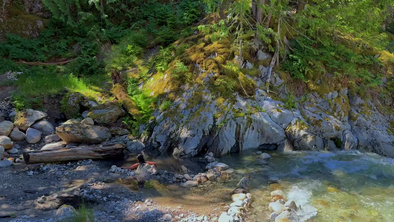 Ripresa in slow motion 4K di una donna che si rilassa nelle sorgenti termali a metà strada, accanto a un impetuoso fiume di montagna, con acqua cristallina, una fitta foresta di conifere e un ambiente selvaggio e pacifico.