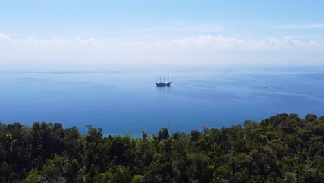 Drones-Aéreos-Que-Se-Elevan-Sobre-Una-Remota-Isla-Tropical-Cubierta-De-Selva-Tropical-Con-Un-Barco-De-Vida-A-Bordo-En-Solitario-En-El-Océano-Azul-Profundo-De-Raja-Ampat,-Papua-Occidental,-Indonesia