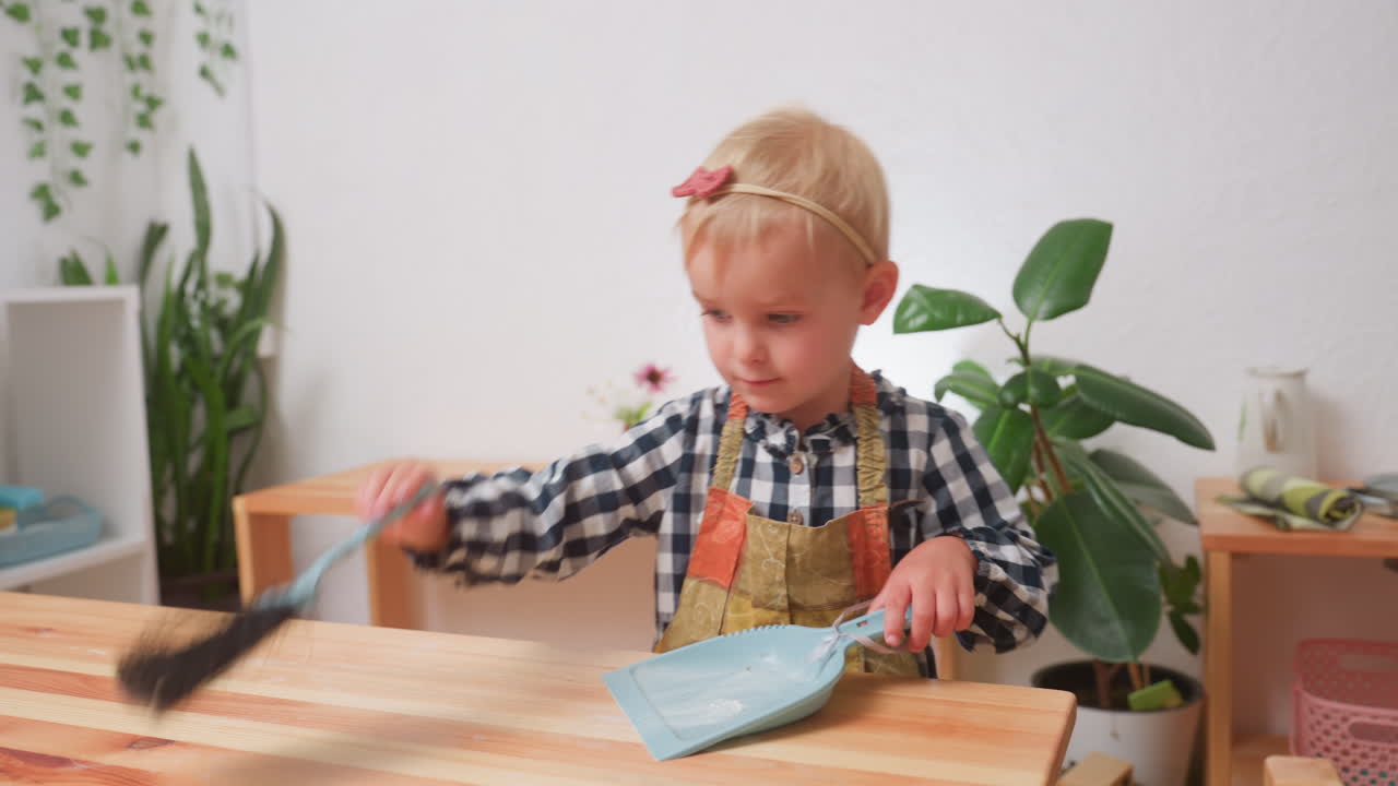 Beautiful child in checkered shirt and apron carefully using brush to sweep flour into blue parker on wooden table inside bright kindergarten with plants, shelves, and utensils